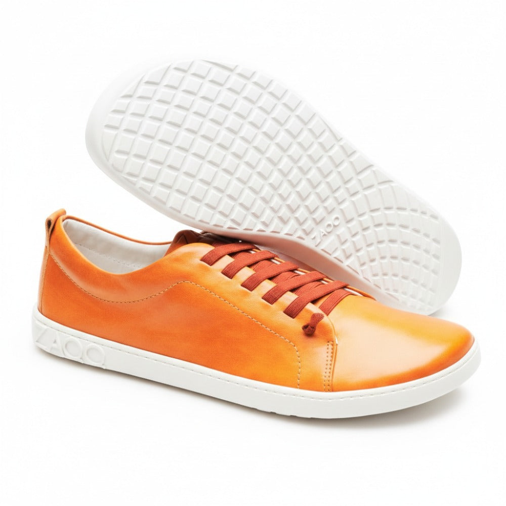 Ausgestellt wird der STIQ Sierra, ein Paar leuchtend orangefarbene Sneaker mit weißer Sohle und roten Schnürsenkeln. Ein Schuh steht aufrecht, während der andere geneigt ist, um die für einen Barfußschuh typische Rautenmustersohle zu präsentieren. Der Markenname ZAQQ ist elegant auf der Seite und der Sohle aus Nappaleder geprägt.